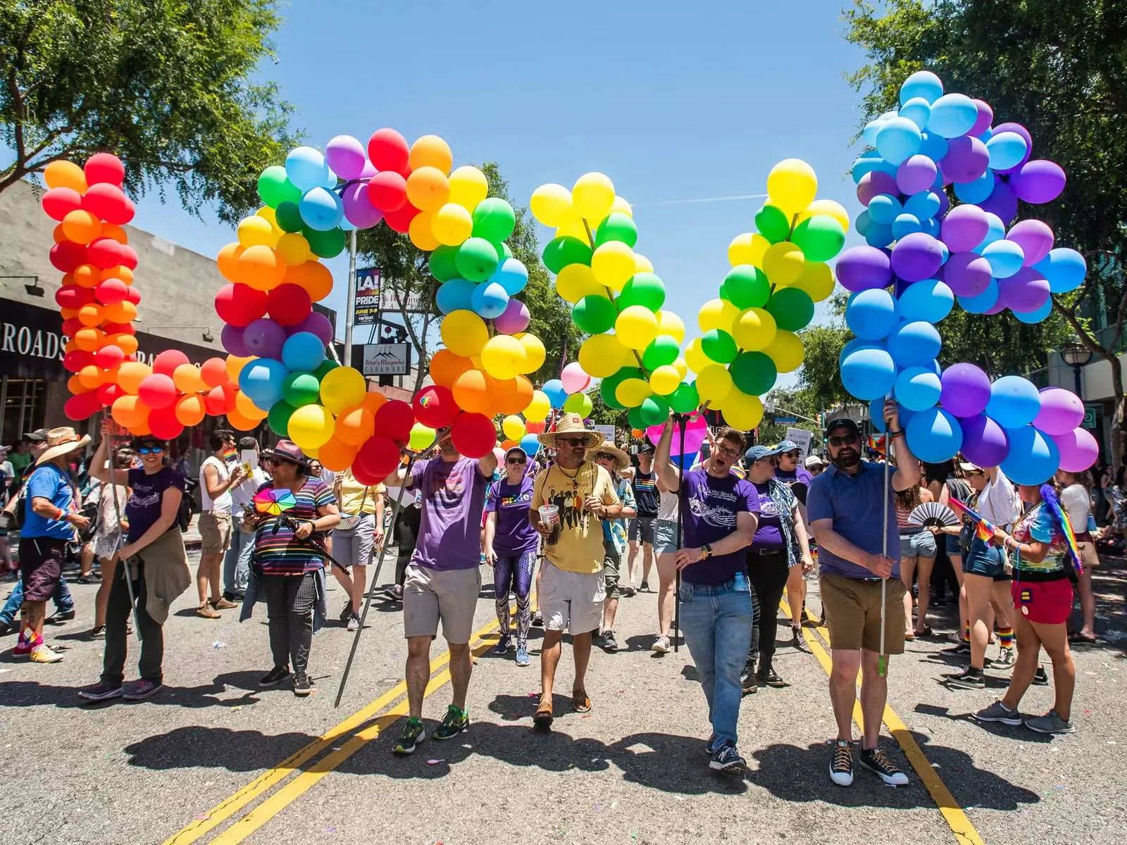 LA Pride marchers carry massive rainbow balloon arches down a sunny street, forming a gateway over the parade route. Marchers at LA Pride carry oversized rainbow balloon arches above the parade route, illustrating one of the biggest Pride celebrations on the West Coast.