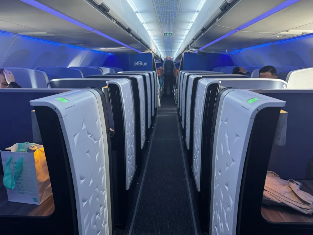 View down the aisle of a JetBlue Mint cabin on an A321XLR, showing rows of suites lit in blue with the jetBlue logo visible at the rear