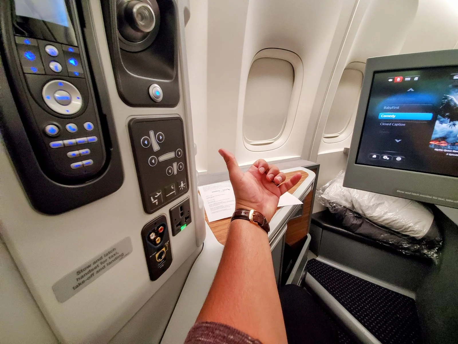 A passenger's arm reaching toward the entertainment screen and seat controls in an American Airlines business class pod.