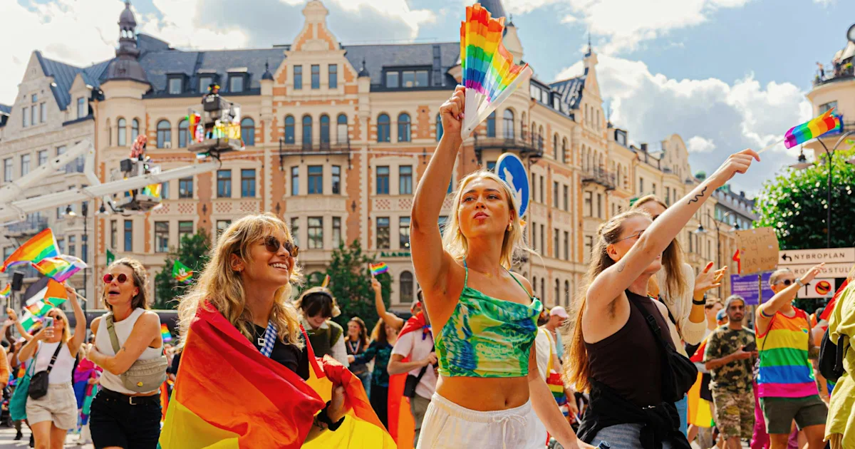 A daytime Pride parade with rainbow flags and a hand-held rainbow fan against European architecture — illustrating the global Pride destinations point.me readers can fly to with points.