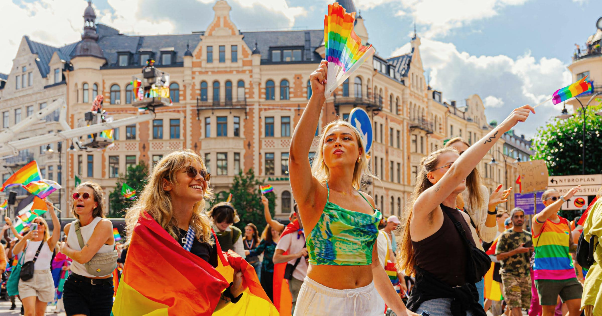 A daytime Pride parade with rainbow flags and a hand-held rainbow fan against European architecture — illustrating the global Pride destinations point.me readers can fly to with points.