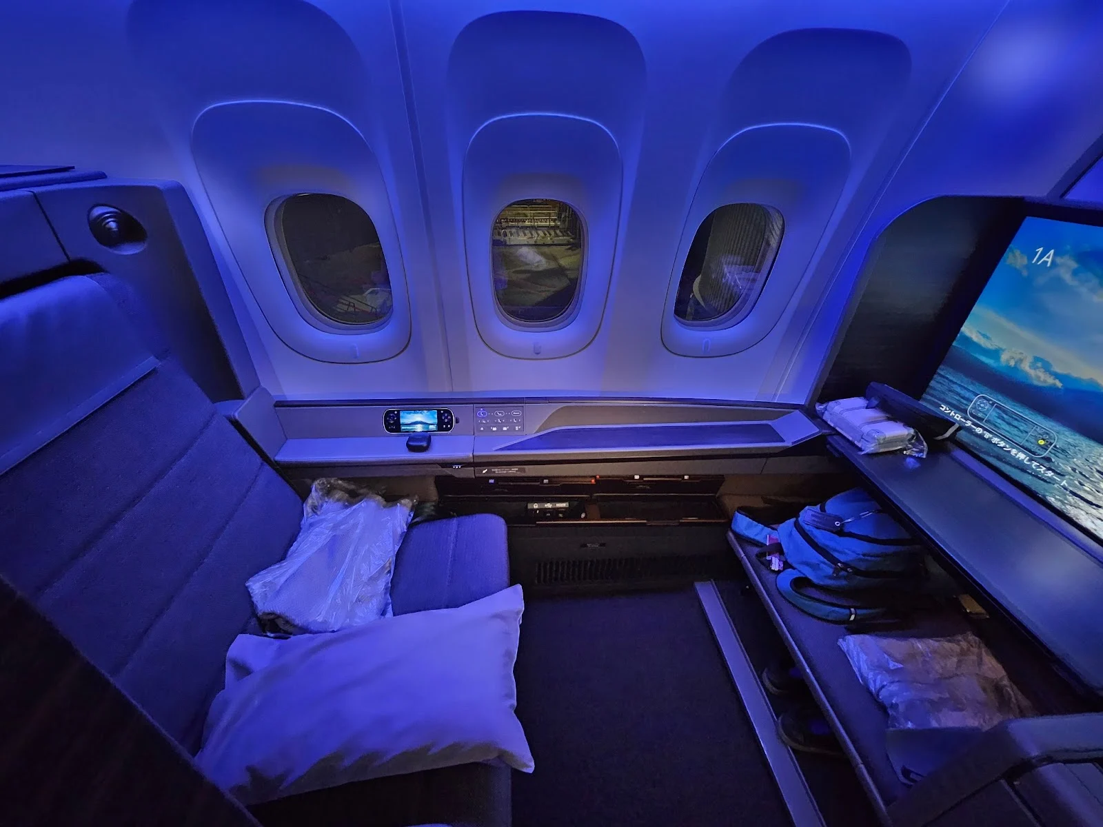 Blue-lit airline premium cabin suite with a lie-flat seat, personal ottoman, and an entertainment screen displaying Japanese-language text