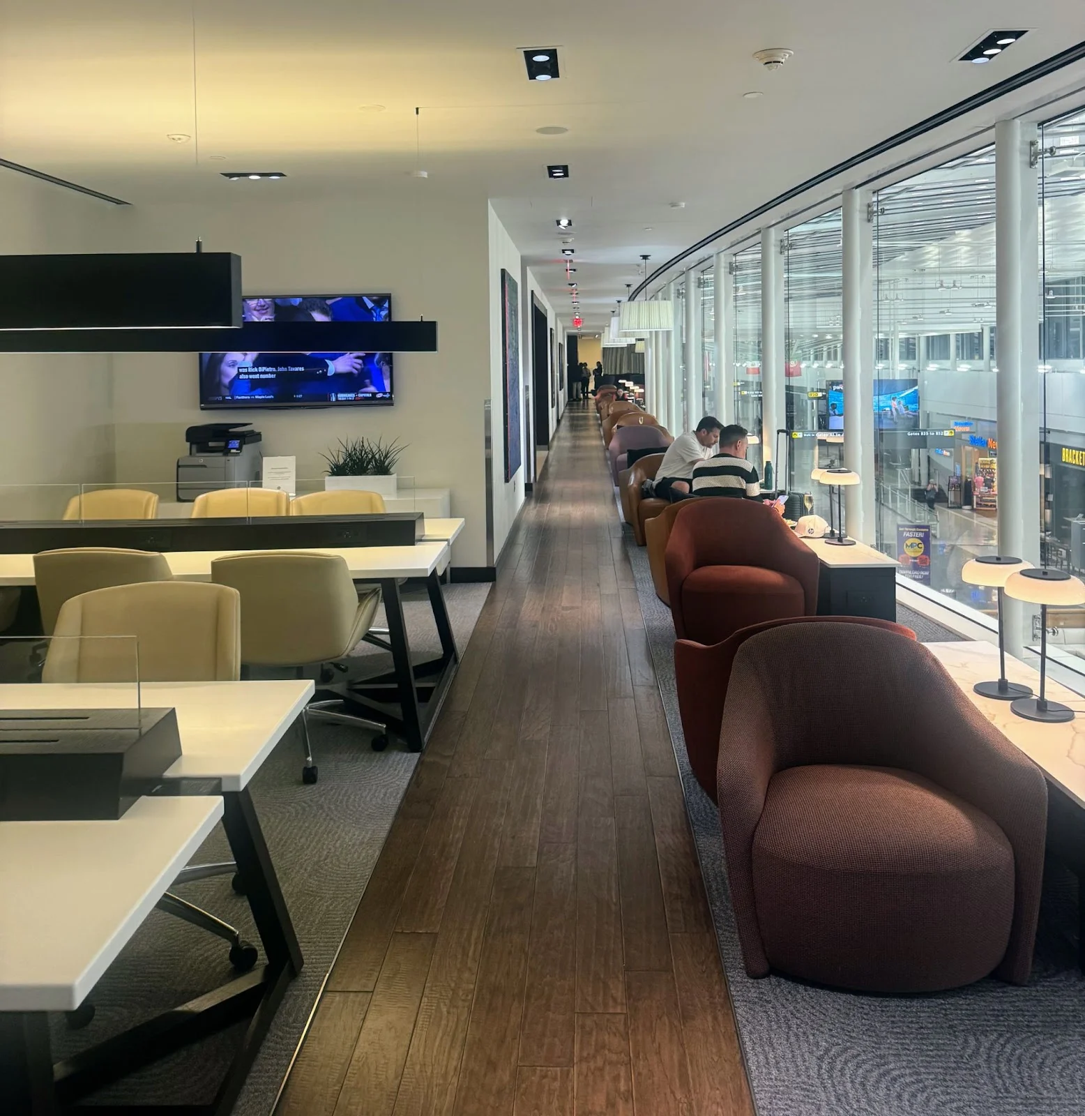 Airport lounge seating area with leather armchairs along a window-lined corridor and a wood-paneled workspace with a TV in the foreground.