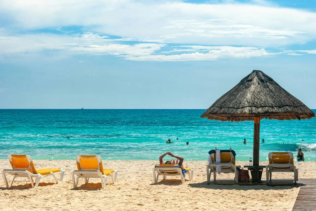 Lounge chairs and a thatched-roof umbrella on a white sand beach in Cancun, with turquoise water and swimmers offshore