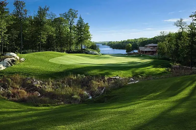 A golf green surrounded by trees and rock outcrops overlooking a lake, with a lodge visible in the distance in Canada