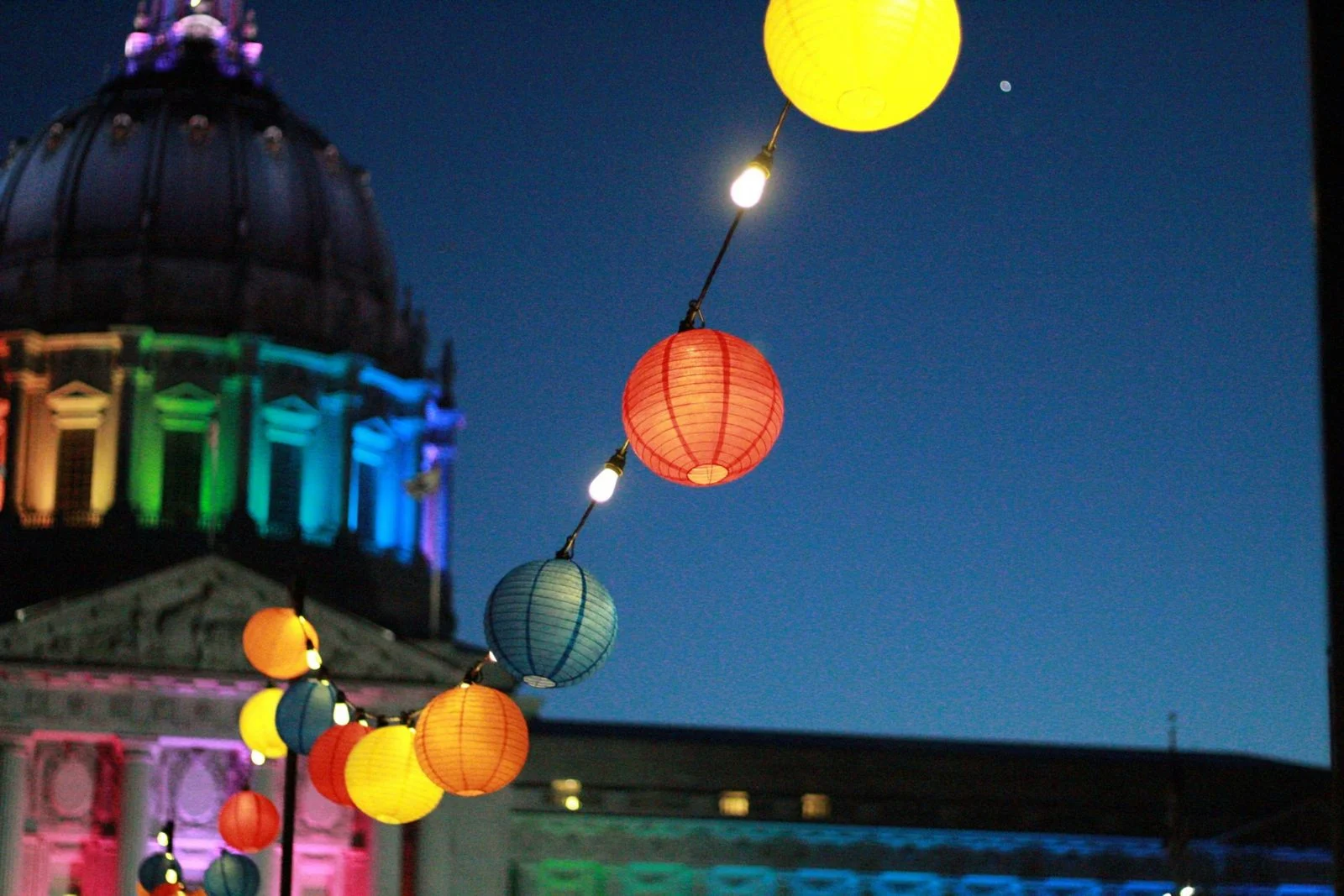 San Francisco City Hall lit in rainbow colors at dusk, with colorful paper lanterns strung across the plaza. San Francisco City Hall lit in rainbow colors at dusk with strings of paper lanterns, marking SF Pride — one of the largest queer celebrations in the U.S.