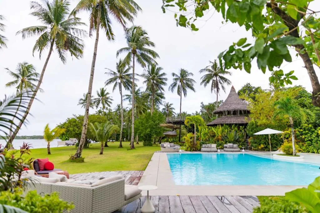 A resort swimming pool surrounded by palm trees and lounge furniture with the ocean visible in the background, Philippines