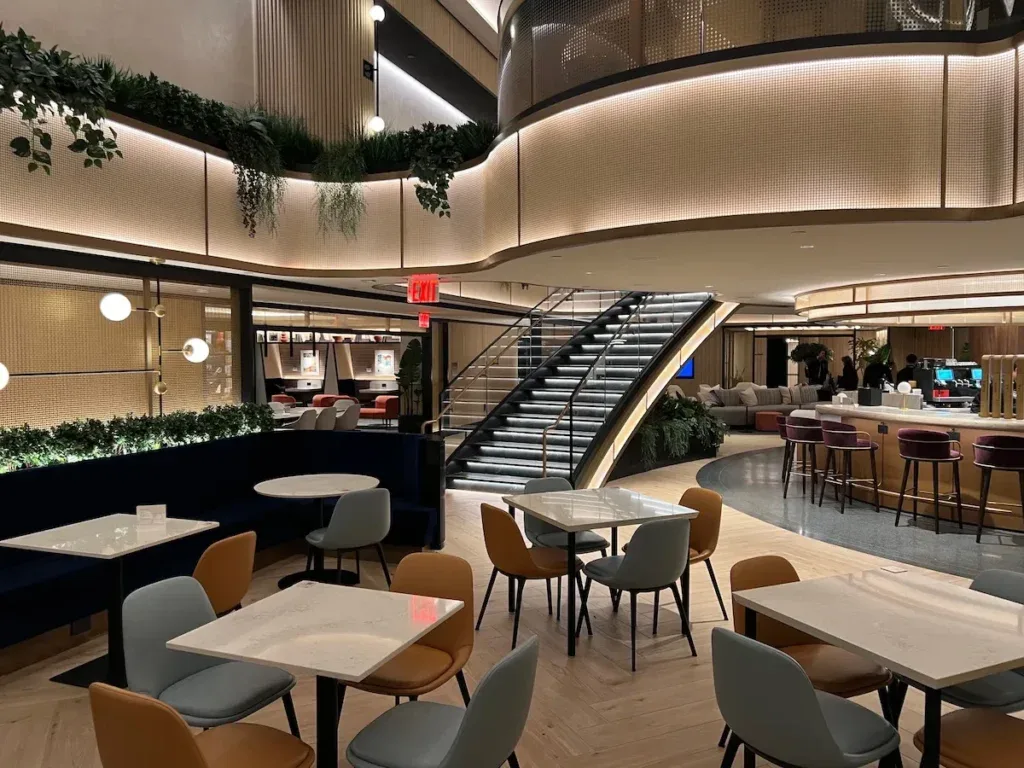 Interior of the Chase Sapphire Lounge at New York LaGuardia Airport, featuring a mezzanine staircase, hanging greenery, and dining tables