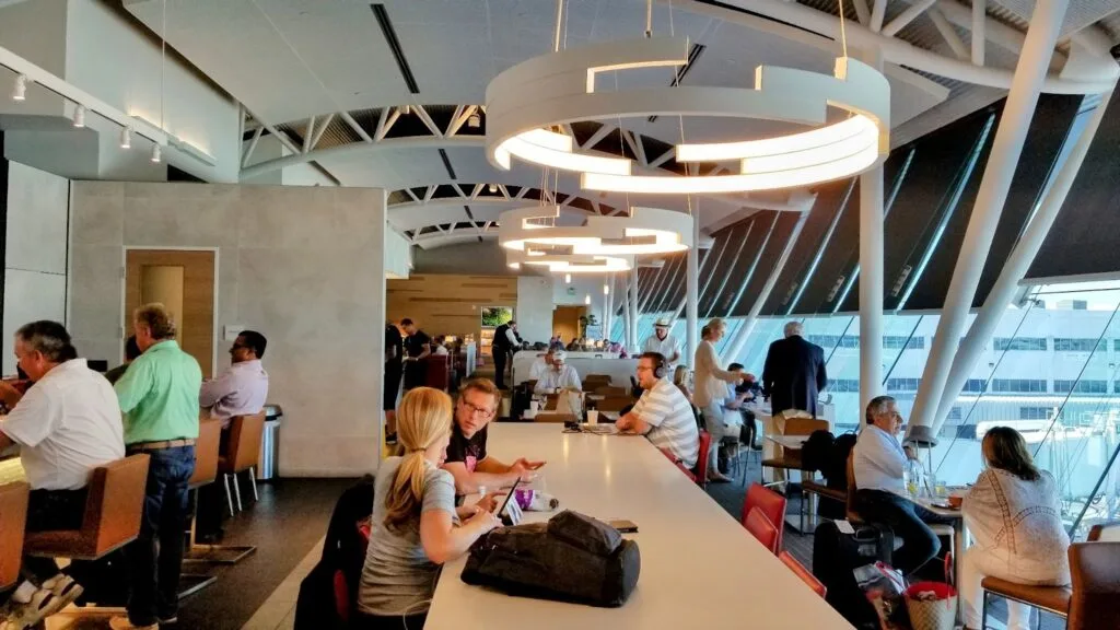 Travelers seated at a communal table in an airport lounge with round pendant lighting and large windows overlooking the tarmac.