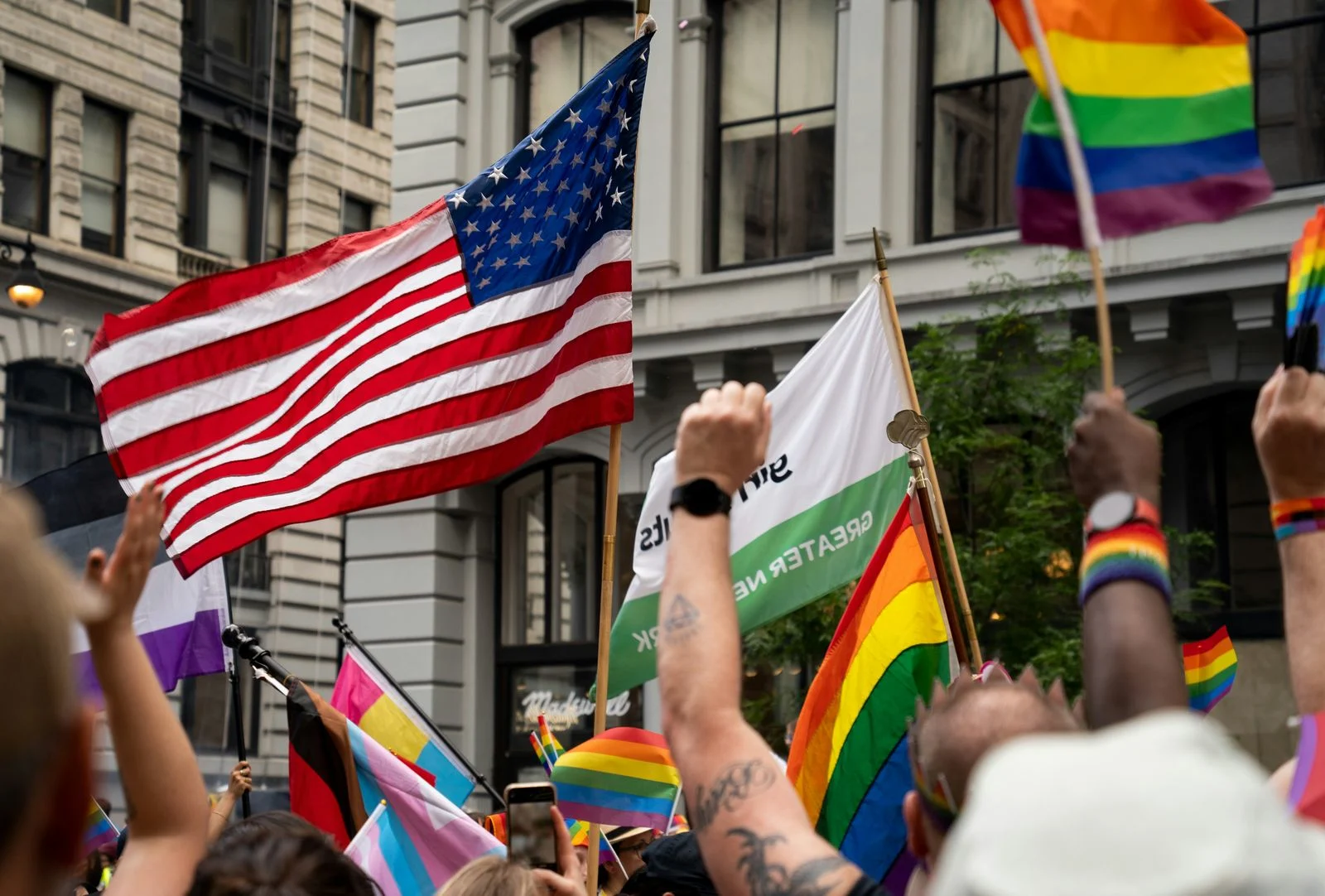 NYC Pride marchers raise an American flag and rainbow flags beneath a banner reading 'Greater New York.' Marchers raise an American flag and rainbow flags at NYC Pride, the parade that traces its origin back to the 1969 Stonewall Uprising.