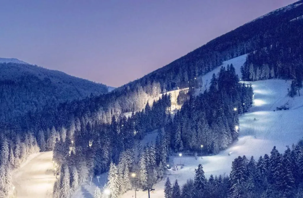 Illuminated ski slopes cut through a snowy forested mountainside at night in Bosnia