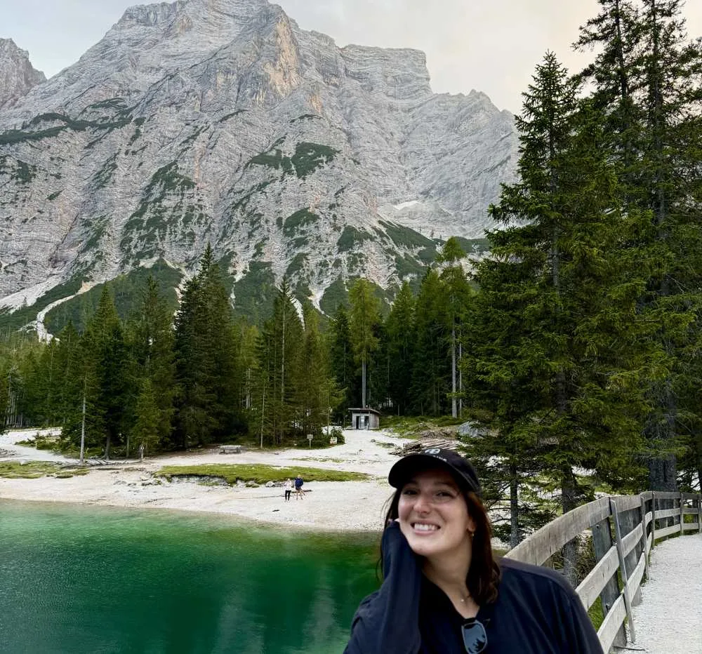 A woman smiling beside a turquoise alpine lake with a rugged mountain peak and pine forest in the background in the Italian Dolomites