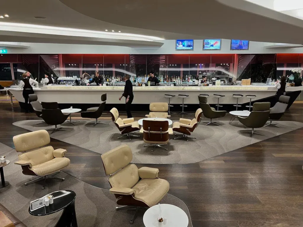 The bar and lounge area of the Virgin Atlantic Clubhouse at London Heathrow, with tan leather Eames lounge chairs and a long marble bar