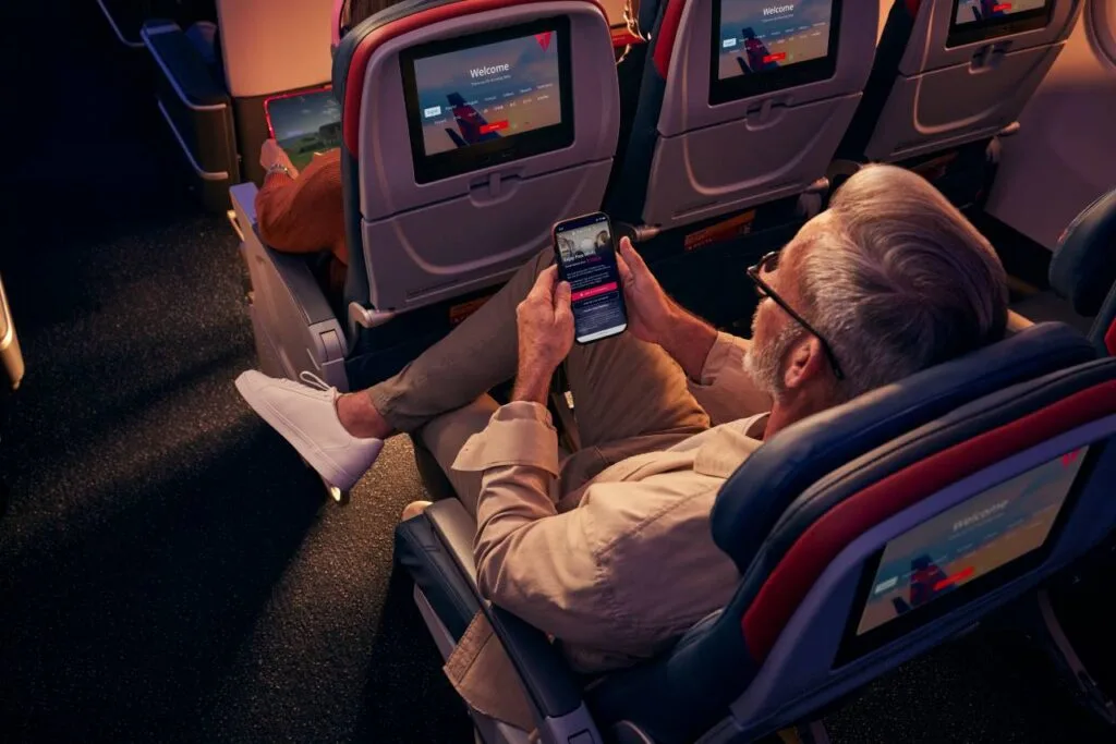 A passenger reclined in a Delta cabin seat at night, using a smartphone next to the seatback entertainment screen.