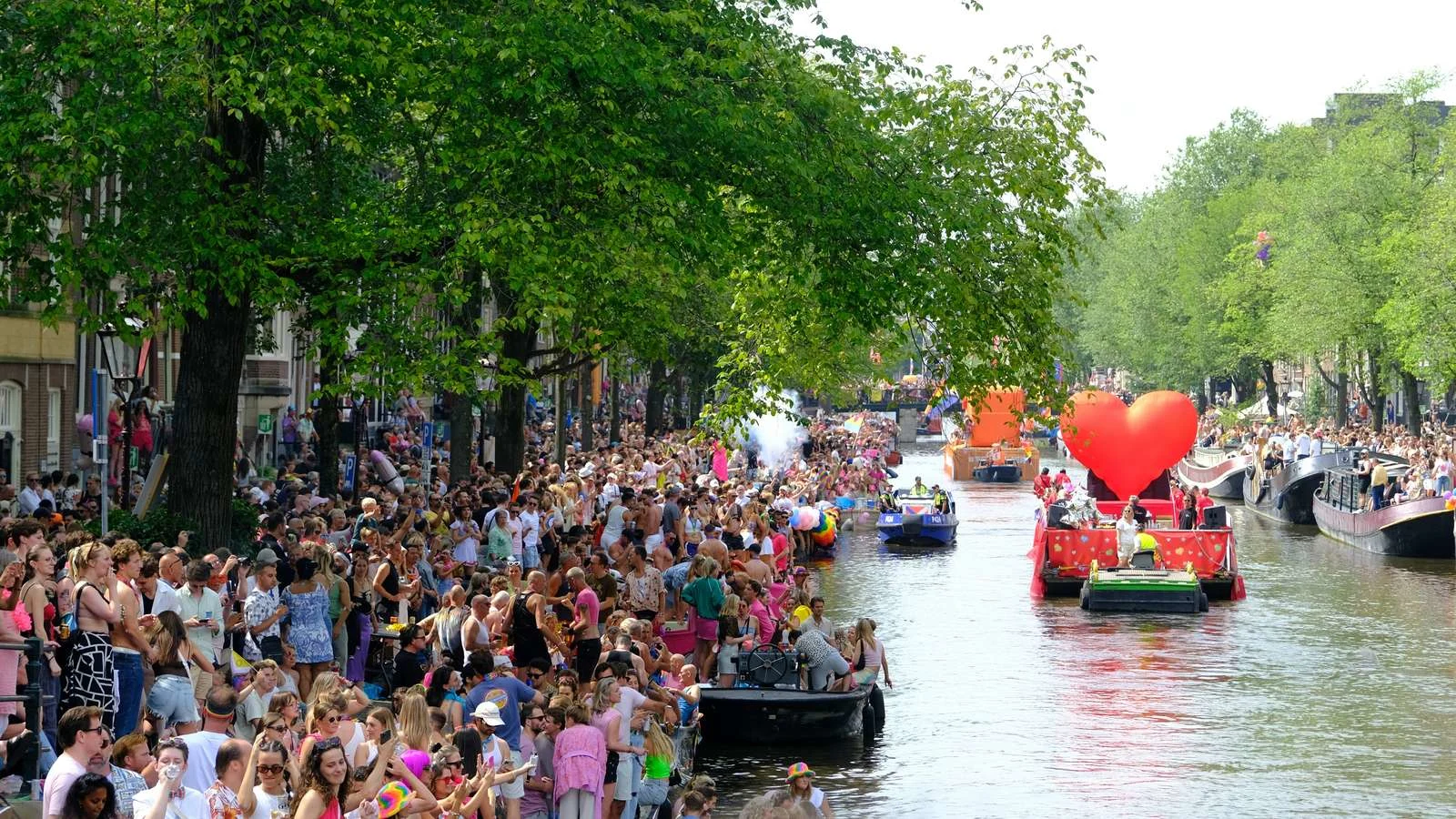 Amsterdam Pride canal parade with crowds lining the banks as a heart-decorated boat passes beneath leafy trees. Amsterdam's iconic Pride canal parade winds between leafy banks of cheering crowds, with a heart-decorated boat at the center — the host city for WorldPride 2026.