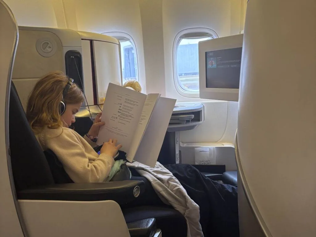 A child wearing headphones reads an in-flight menu while seated in a business class seat with a personal entertainment screen nearby.