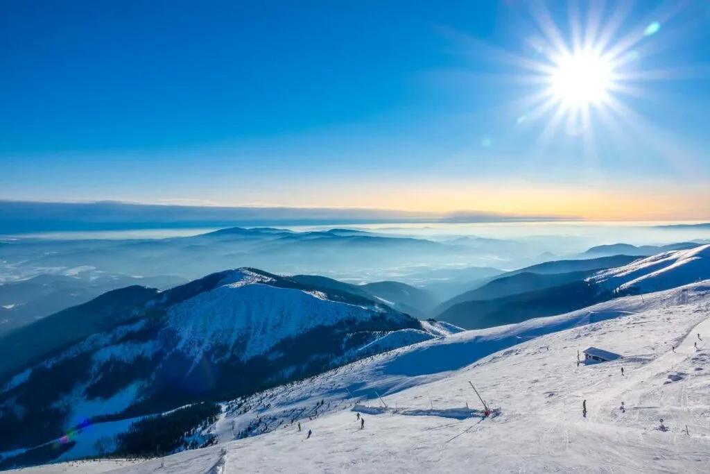 A sun-drenched view over layered mountain ranges and a snowy ski slope in Slovakia, with skiers visible below