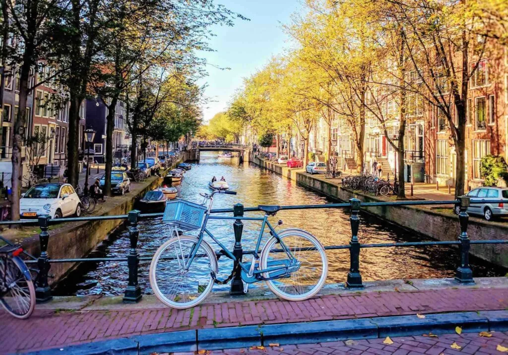 A bicycle parked on a bridge railing overlooking an Amsterdam canal lined with houseboats, trees, and canal houses