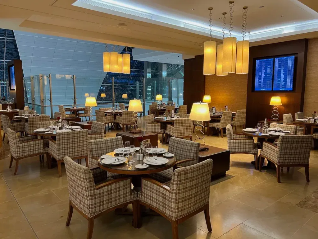 Dining area of the Emirates First Class Lounge in Dubai, with plaid armchairs at round tables and a flight information screen in the background
