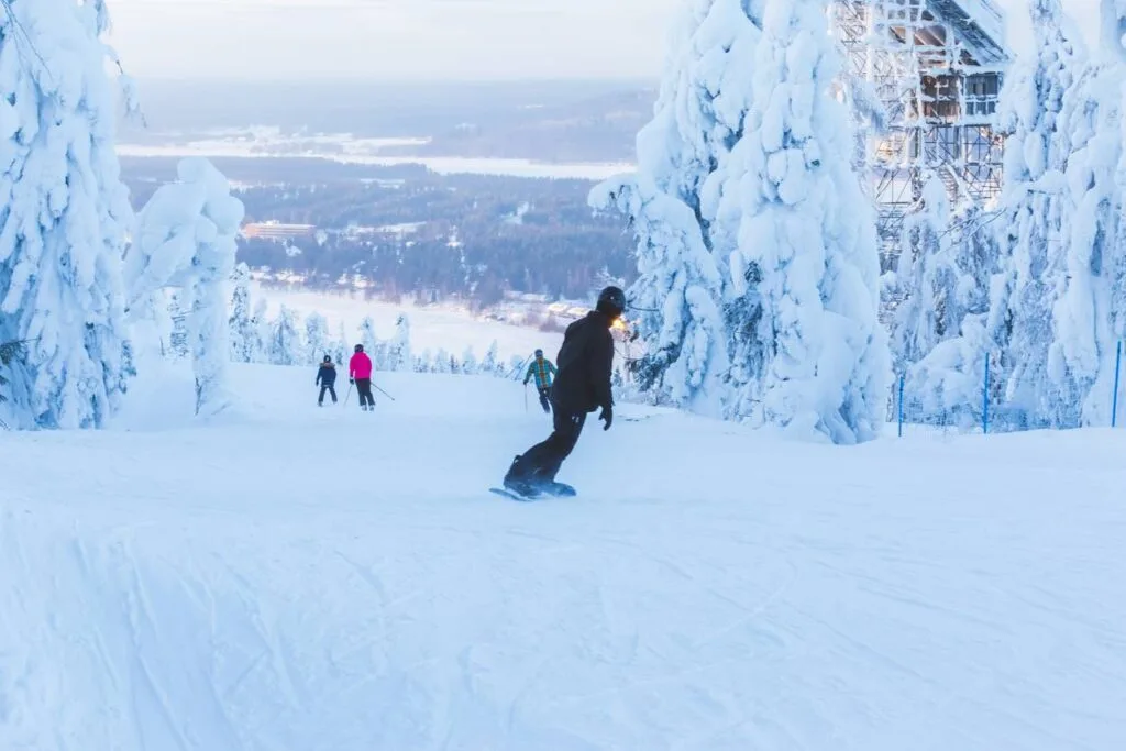 A snowboarder descends a slope in Finland past frost-covered trees, with a ski jump tower visible in the background