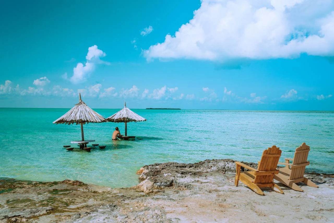 Thatched-roof umbrellas standing in shallow turquoise water off a rocky Belize shoreline, with wooden chairs on the rocks