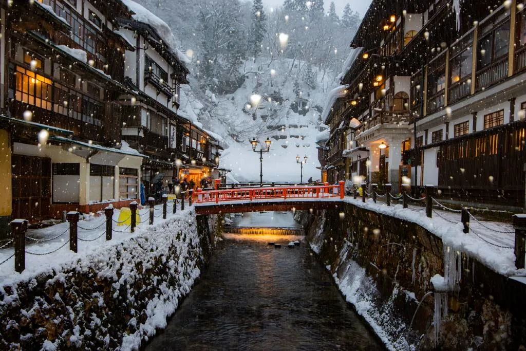 Snow falling over the illuminated ryokans and red bridge along the canal in Ginzan Onsen, Japan