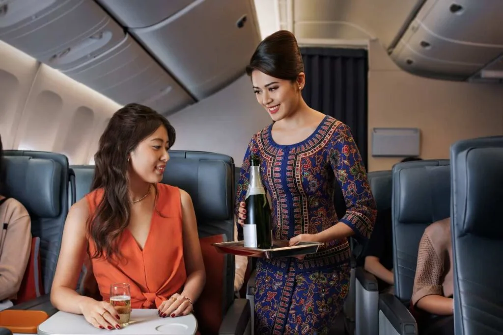 A Singapore Airlines flight attendant in a traditional batik uniform serves champagne to a passenger in premium economy.