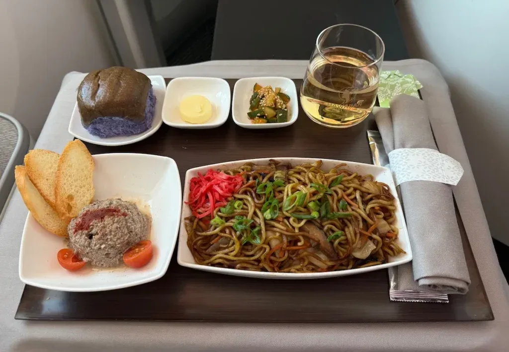 A Hawaiian Airlines first class meal tray featuring noodles, a tomato appetizer, taro bread, and a glass of white wine