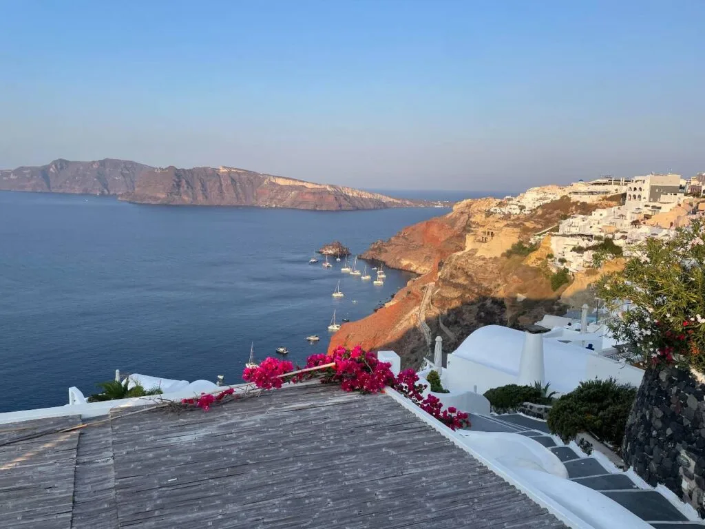 View from a Santorini cliffside terrace with bougainvillea in bloom, overlooking whitewashed buildings, the caldera, and sailboats at sunset