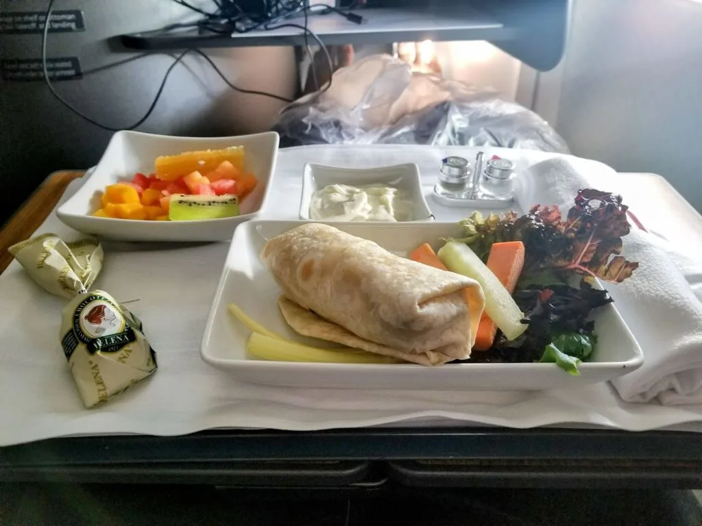 An American Airlines business class meal tray with a wrap, fresh vegetables, fruit, and a cheese wedge.