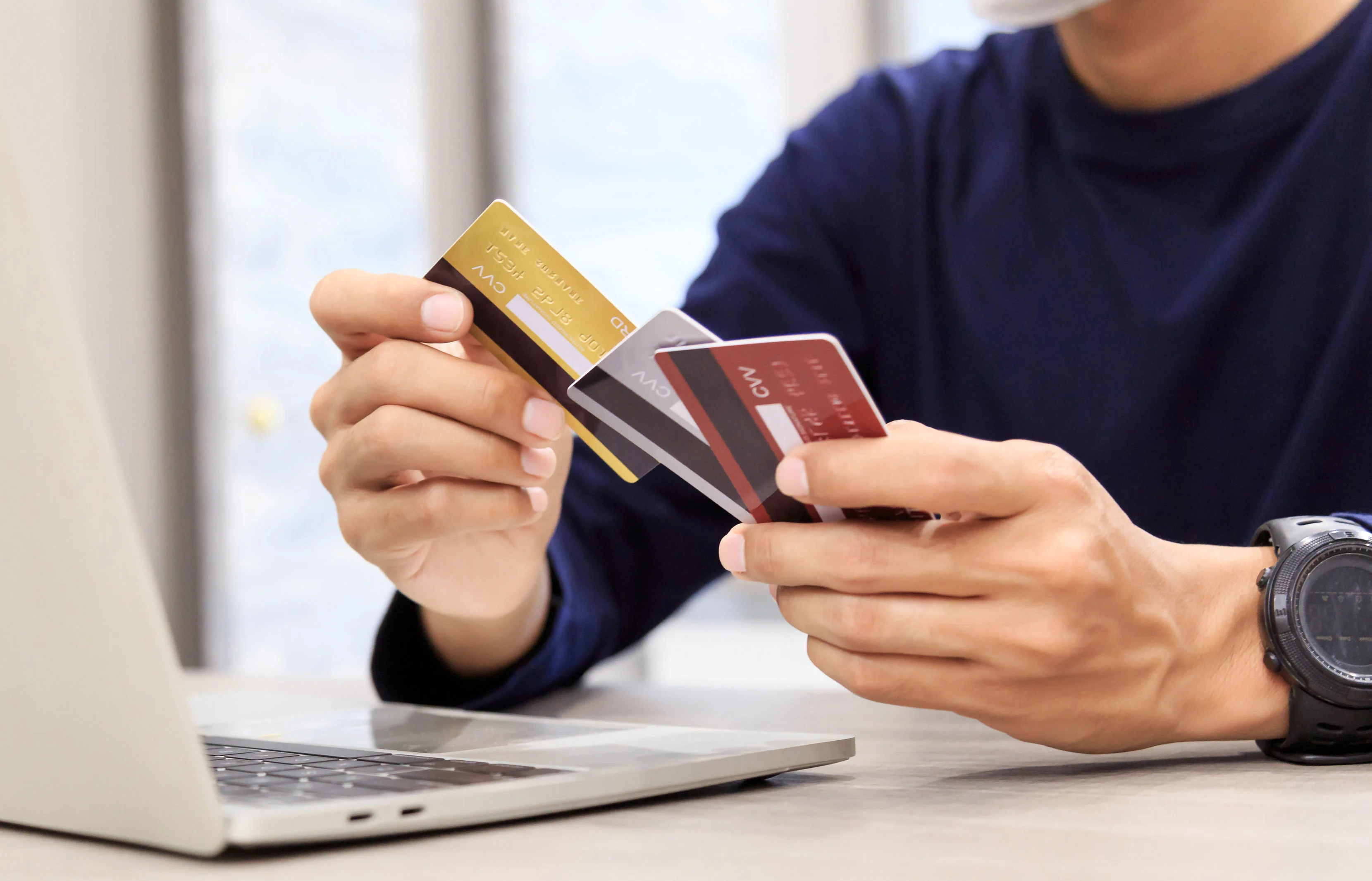 Close-up of a person's hands holding three credit cards while sitting at an open laptop, comparing which card to use.