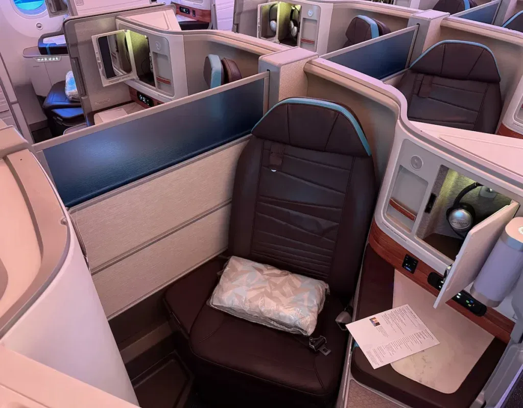 A Hawaiian Airlines first class suite aboard a Boeing 787, featuring a private brown leather seat with a pillow and blue accent trim