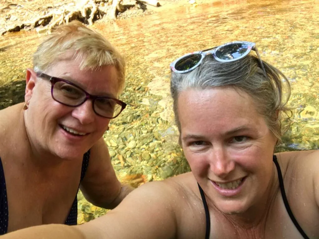 Two women smiling for a selfie while swimming in a clear, sunlit creek