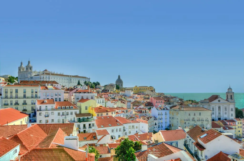 Aerial view of Lisbon's Alfama district with terracotta rooftops, historic church domes, and the river in the distance under a clear blue sky.
