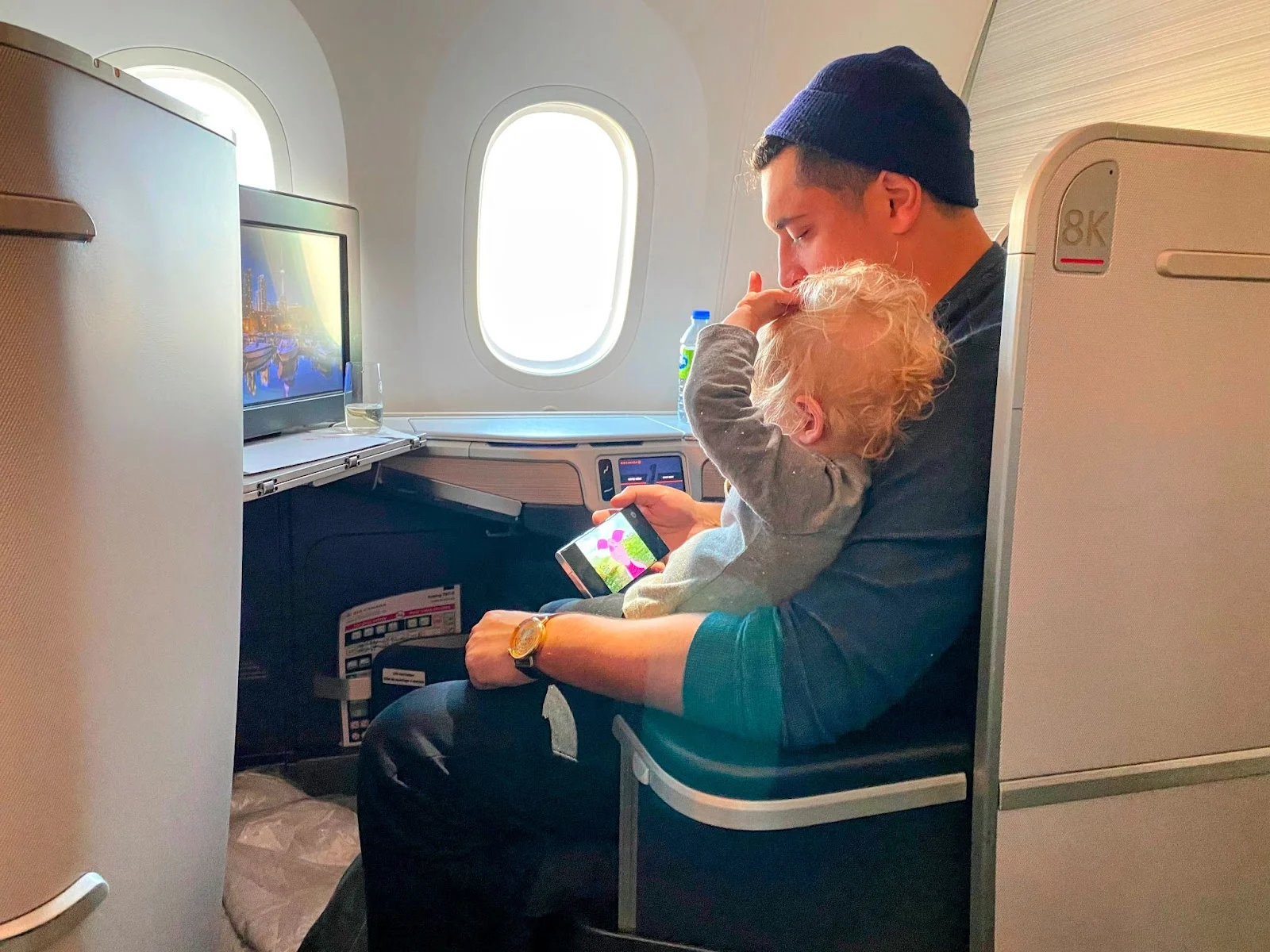 A father holds a toddler while showing her a phone video in an Air Canada business class seat, with the entertainment screen playing in the background.