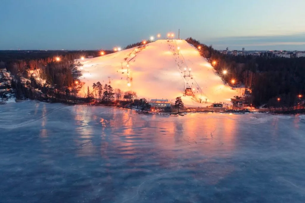 An illuminated ski slope glows at dusk beside a frozen lake in Sweden, with skiers visible on the run