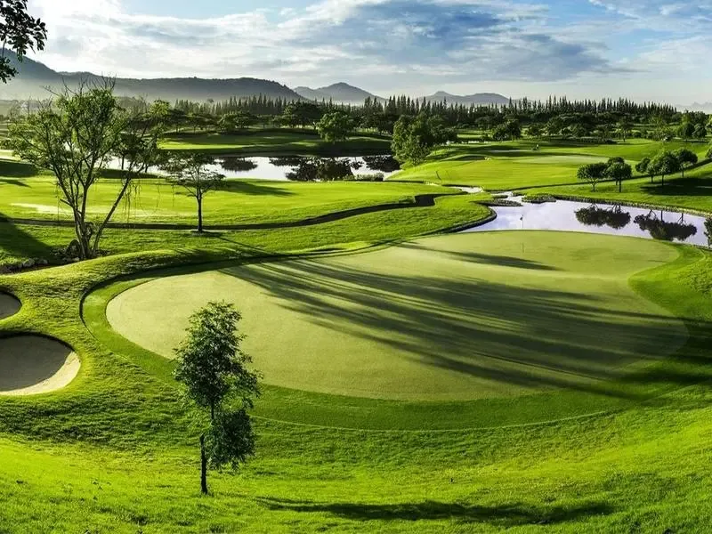 A lush green golf course with a water hazard and sand bunkers, framed by trees and distant mountains in Thailand