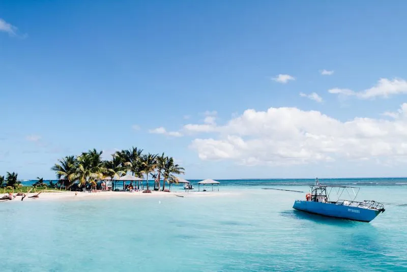 A picturesque tropical paradise featuring a small sandy island with palm trees and beach umbrellas in the center. The island is surrounded by crystal-clear turquoise waters. A blue boat is anchored in the shallow lagoon to the right side of the image. The scene is set under a bright blue sky with scattered white clouds. This idyllic destination appears to be a small cay or sandbar that serves as a day trip spot for tourists in the Caribbean or similar tropical region.