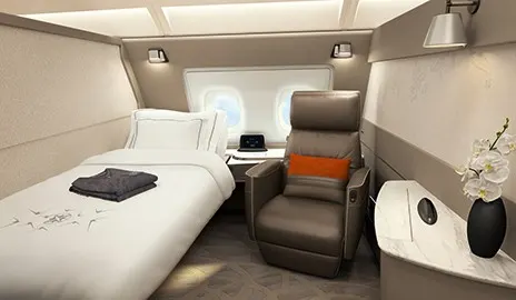 A private airline suite aboard an A380 featuring a separate armchair and a made-up bed with white linens, reading lights, and a small vanity area with flowers