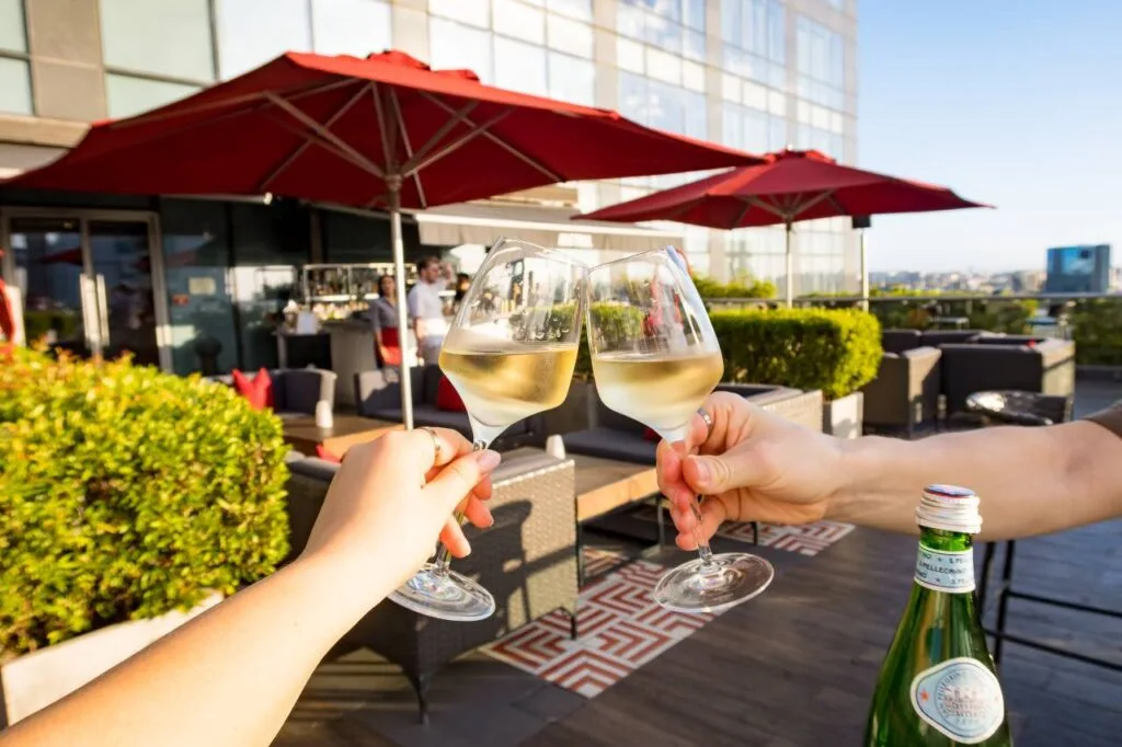 Two people clinking wine glasses together on a rooftop terrace with red umbrellas and a city skyline in the background