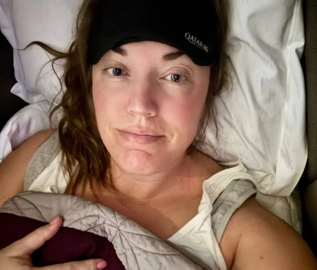 A woman wearing a Qatar Airways sleep mask smiles for a selfie while lying in a Qsuite business class bed