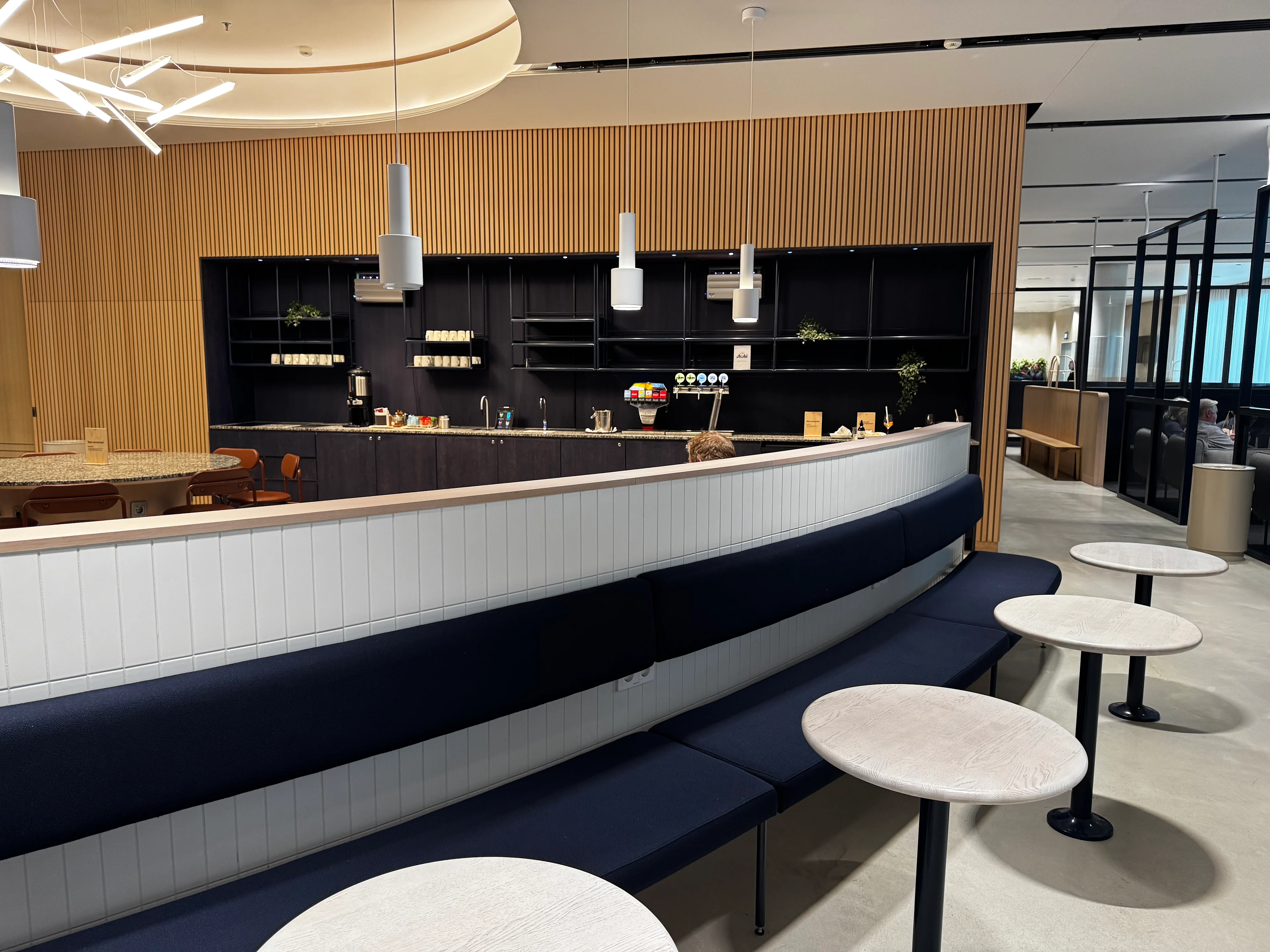 Finnair lounge seating area in Helsinki Airport with a curved navy banquette, round cocktail tables, and a self-serve bar in the background.