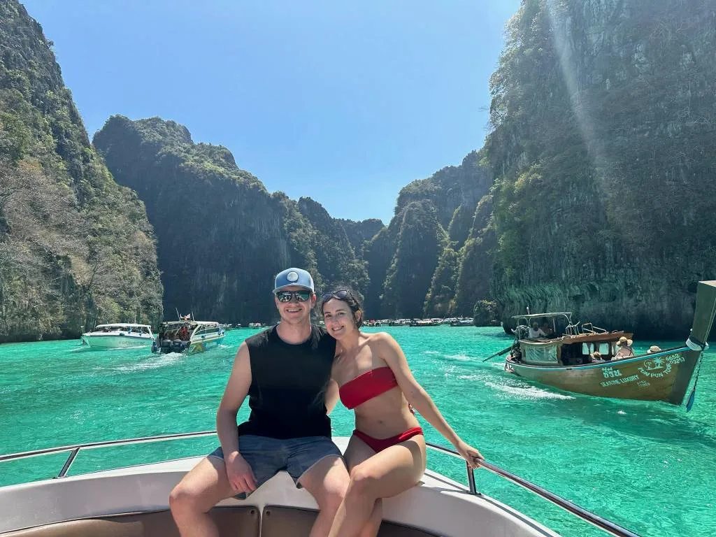A couple sitting on the bow of a boat in turquoise water surrounded by limestone cliffs, with longtail boats nearby.