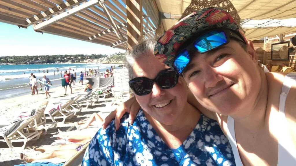 Two women smiling in a beachside selfie with sun loungers and the ocean in the background.