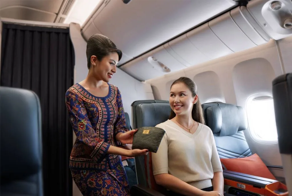 A Singapore Airlines flight attendant in a traditional sarong kebaya uniform hands an amenity kit to a premium economy passenger