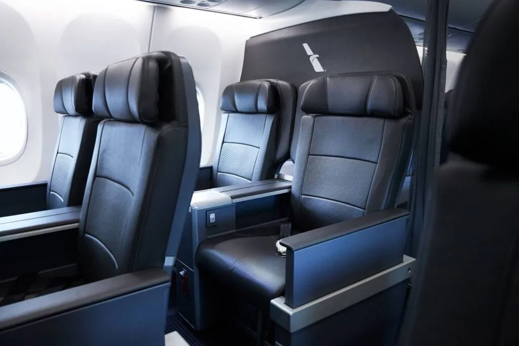 Black leather recliner seats in a domestic first class cabin, with a small airline logo on the headrest covers and a fold-out tray table between seats