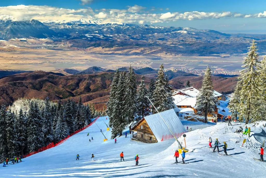 Skiers pass a wooden mountain chalet on a ski slope overlooking the Carpathian foothills in Romania