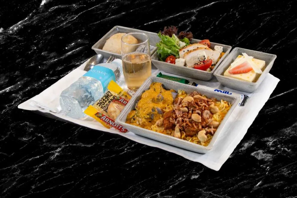 Singapore Airlines premium economy meal tray with lamb biryani, salad, bread roll, cheese plate, and a glass of champagne.