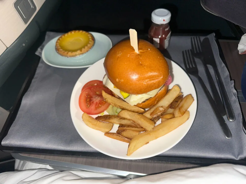 A burger and fries with a tomato slice and a small tart, served on a Cathay Pacific business class tray table