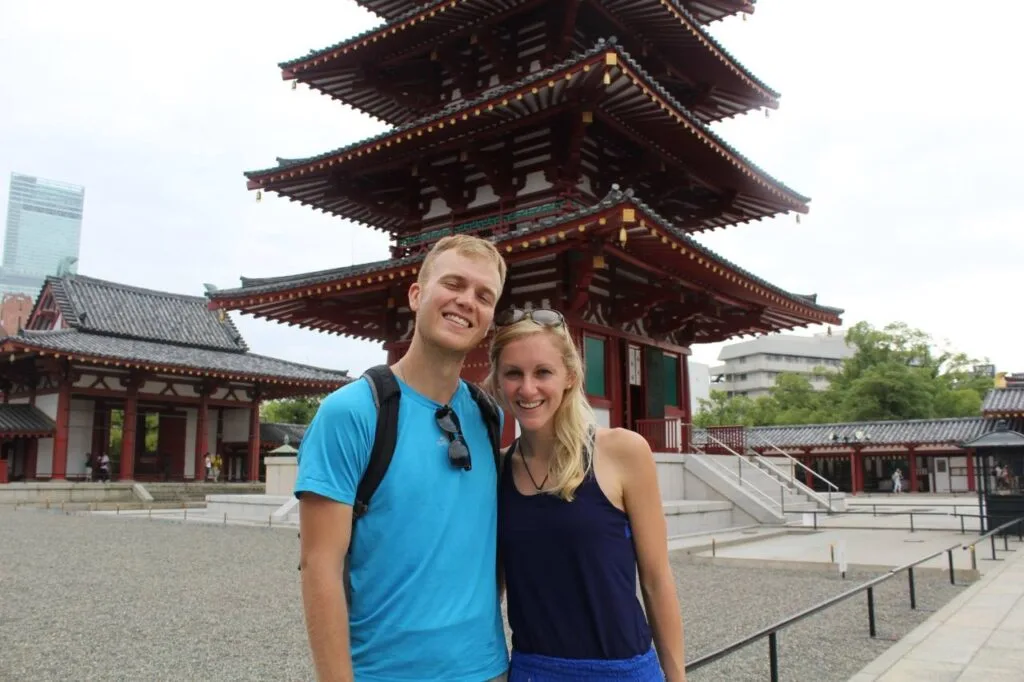 A couple posing together in front of a red multi-tiered pagoda at a Buddhist temple in Japan.